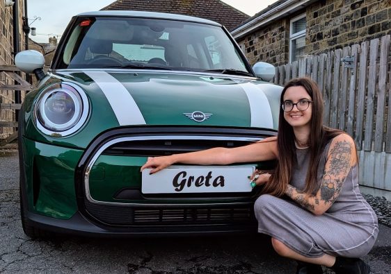 Emma perched in front of her new green mini cooper with white stripes on the bonnet, and white wing mirrors and roof. In front of the car, she is holding a temporary fake reg plate reading 'Greta' the affectionate name of her car. Emma has a sleeve of tattoos and is wearing a grey midi dress.