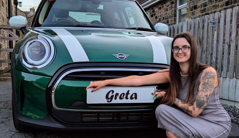 Emma perched in front of her new green mini cooper with white stripes on the bonnet, and white wing mirrors and roof. In front of the car, she is holding a temporary fake reg plate reading 'Greta' the affectionate name of her car. Emma has a sleeve of tattoos and is wearing a grey midi dress.